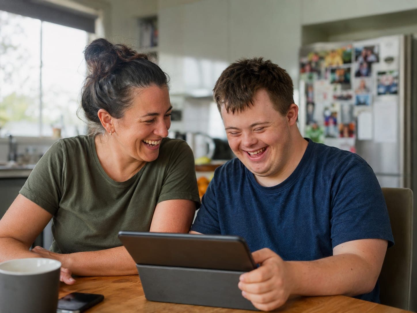A support worker and a young man laughing together over a tablet at a kitchen table